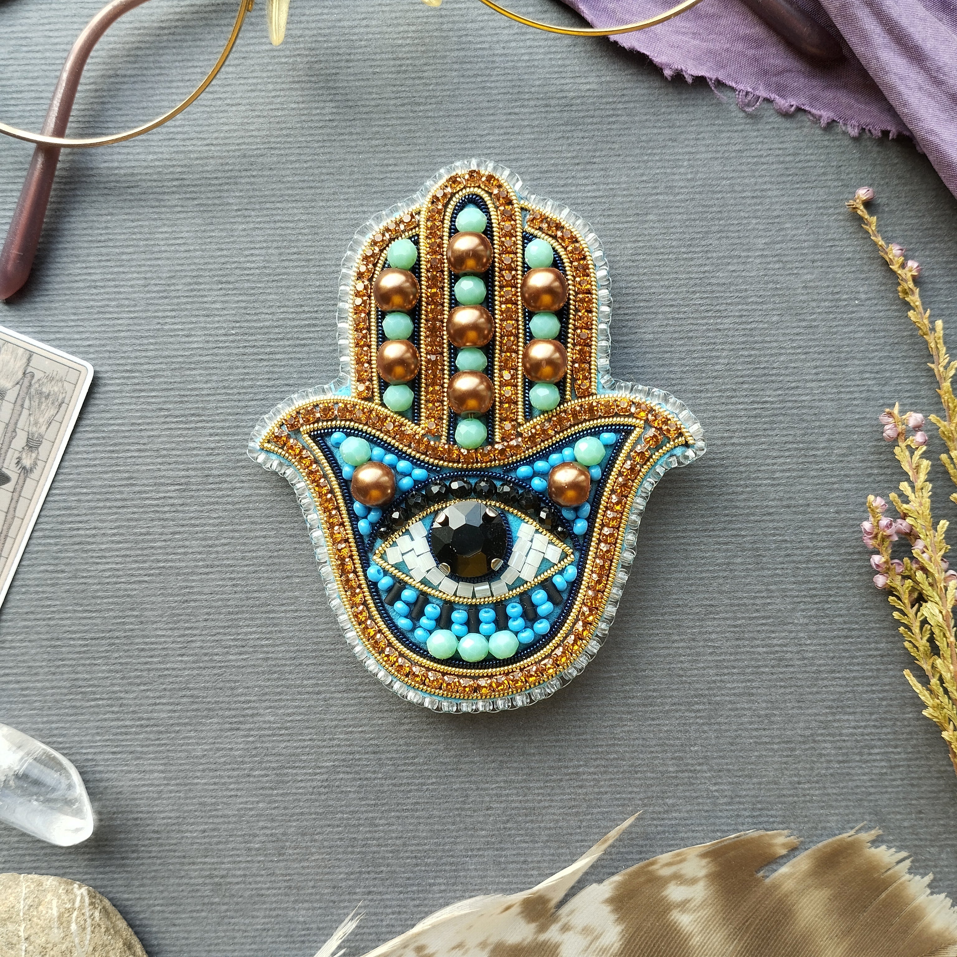 a handcrafted piece of jewelry, a hamsa charm, adorned with various colorful beads and gemstones. The charm is placed on a gray surface, surrounded by other objects such as feathers, crystals, and a card.